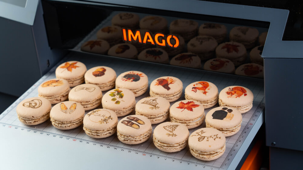 Kolorowy druk spożywczy na makaronikach za pomocą specjalistycznego urządzenia drukującego, ploter cukierniczy Imago Vulpes. Na obrazie widać serię jasnych makaroników ozdobionych precyzyjnymi, jadalnymi obrazami kwiatów i wzorów.